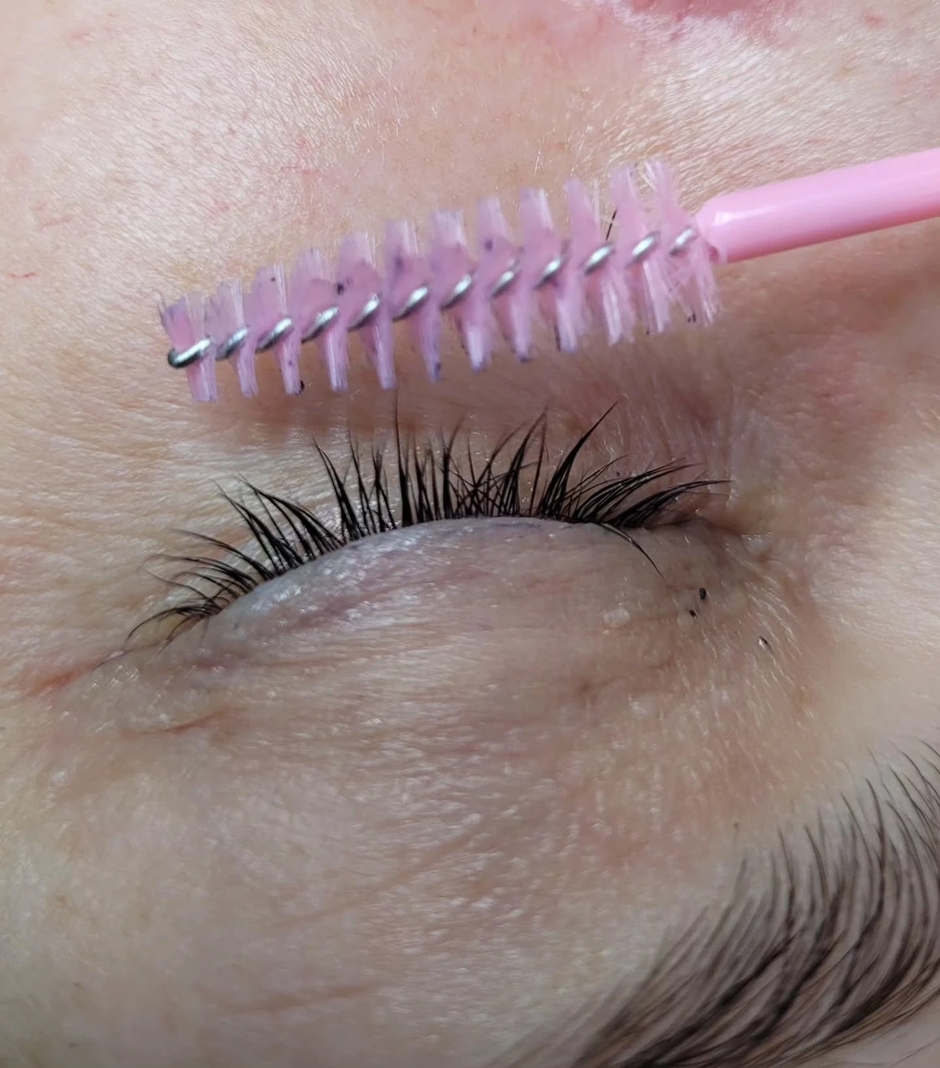 Marina Litwin - Behandlungen für Gesicht, Körper, Hände und Füße - Nahaufnahme eines geschlossenen Auges mit langen, dunklen Wimpern. Ein pinkfarbenes Mascarabürstchen wird knapp über dem Lid gehalten, bereit, die Wimpern zu tuschen.