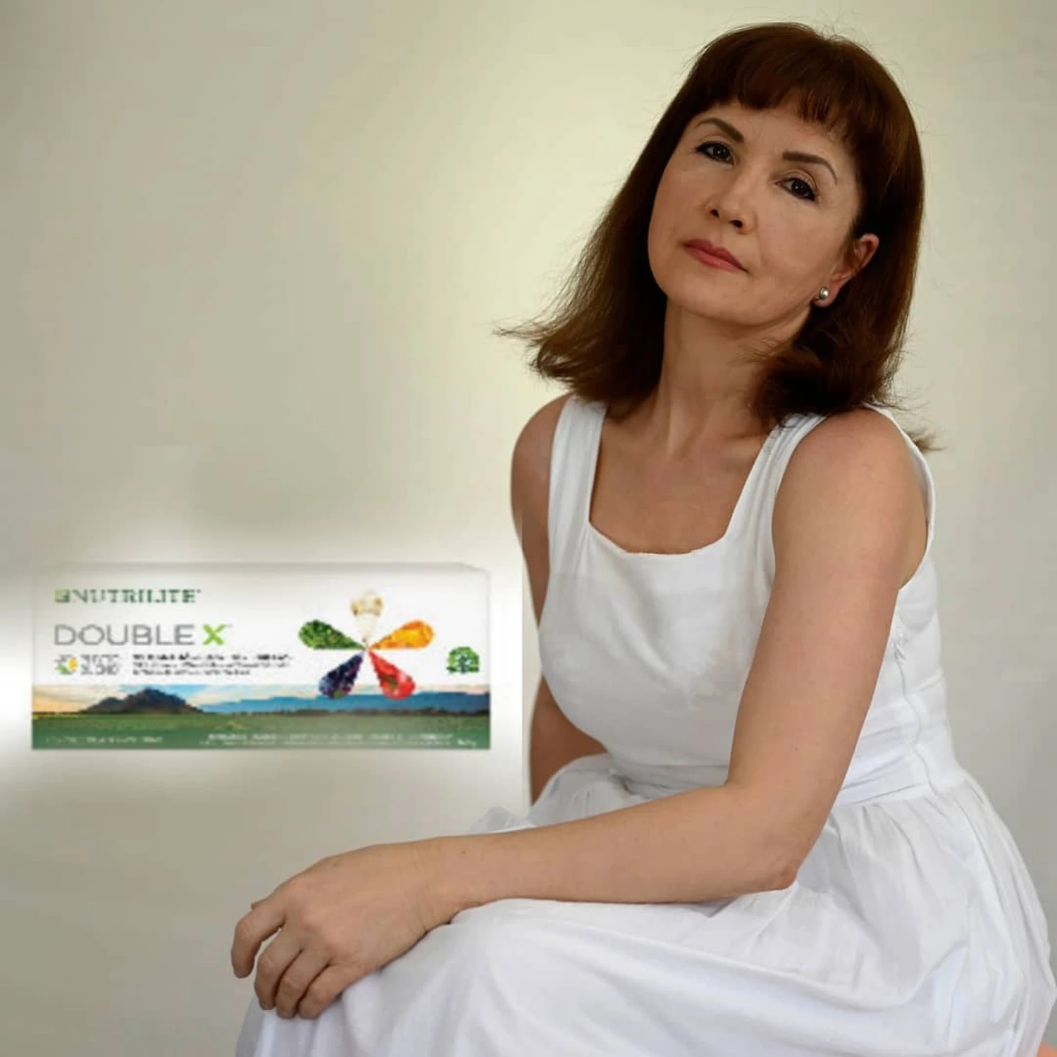 Marina Litwin - Behandlungen für Gesicht, Körper, Hände und Füße - Eine ältere Frau mit schulterlangem braunem Haar und Pony sitzt in einem weißen ärmellosen Kleid und posiert neben einer Packung Nutrilite Double X Multivitaminpräparate.