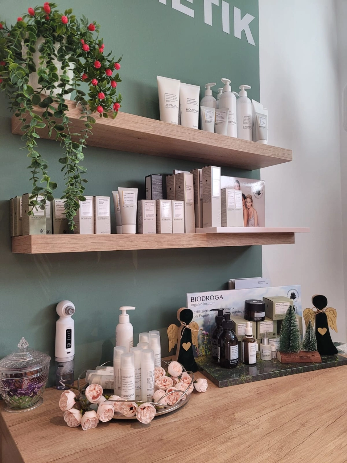 Marina Litwin - Behandlungen für Gesicht, Körper, Hände und Füße - Eine minimalistische Präsentation von Hautpflegeprodukten auf hellen, schwebenden Holzregalen vor einer dunkelgrünen Wand, mit einer hängenden Topfpflanze mit roten Beeren und Dekorationselementen wie Rosen und kleinen Figuren auf einer Holztheke darunter.