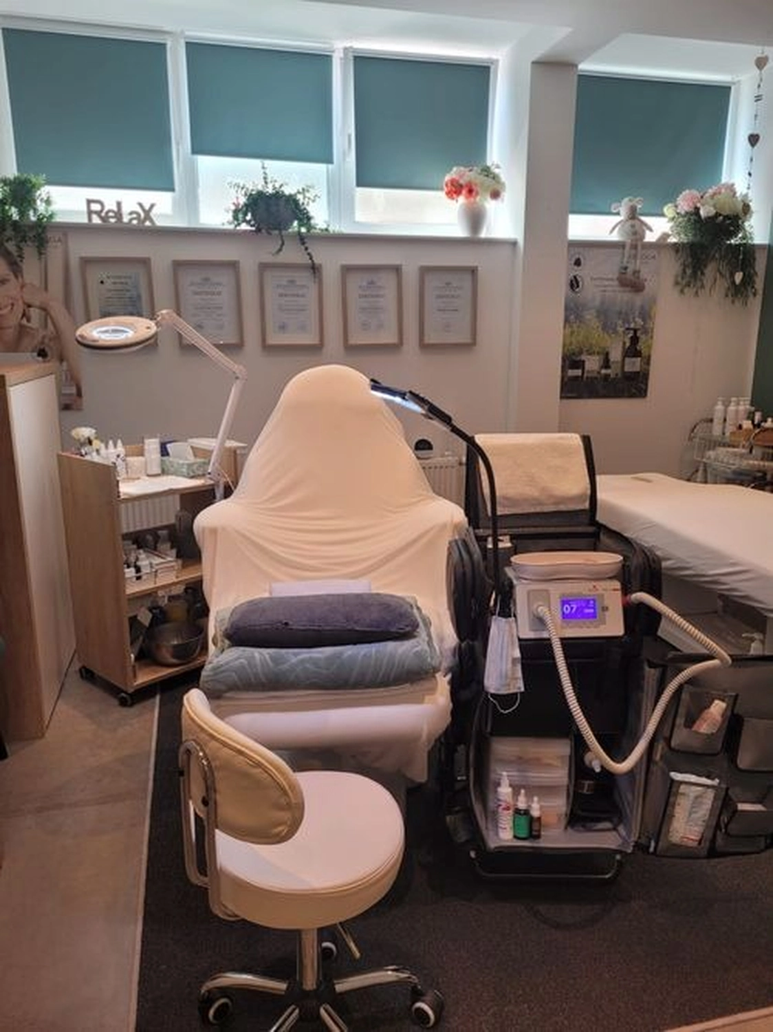 Marina Litwin - Behandlungen für Gesicht, Körper, Hände und Füße - Ein Behandlungsraum in einem Spa oder einer ästhetischen Klinik mit einem mit weißem Stoff bezogenen Liegesessel, professionellen Hautpflegegeräten auf einem Wagen, einem Rollhocker und mehreren gerahmten Zertifikaten an der Wand.